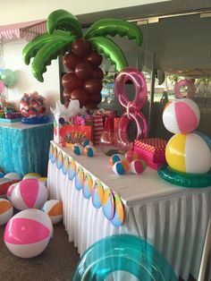 a table topped with lots of balloons and beach balls next to a palm tree on top of a white table cloth