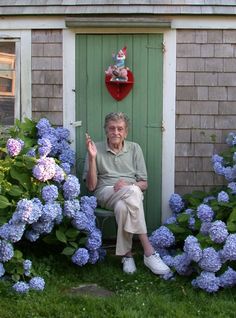 an older man sitting in front of a green door surrounded by blue hydrangeas