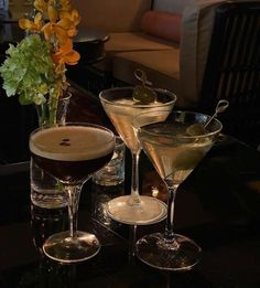 three martini glasses sitting on top of a table next to a vase filled with flowers