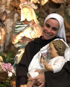 a woman holding a doll in front of a wall with statues and flowers on it