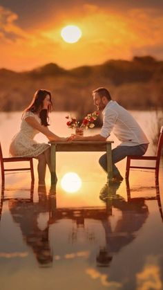 a man and woman sitting at a table in front of a lake with the sun setting