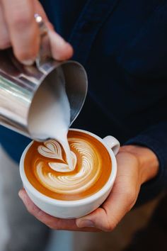 a person pouring milk into a cup of latte artisan style coffee in their hands