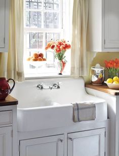 a white kitchen sink sitting under a window next to a bowl of fruit and flowers