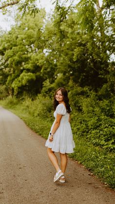 a girl in a white dress standing on the side of a road with trees and bushes behind her