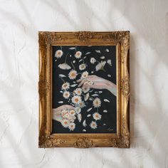 a painting hanging on the wall with white flowers in it's gold frame and hands reaching for daisies