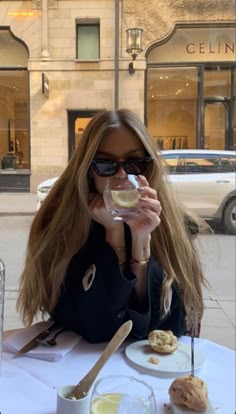 a woman sitting at a table drinking from a wine glass in front of a building