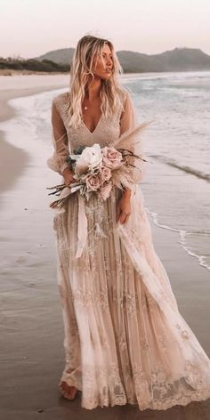 a woman is standing on the beach wearing a long dress with flowers in her hand