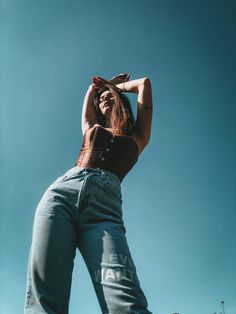 a woman in high waisted jeans is standing with her hands behind her head and looking up into the sky