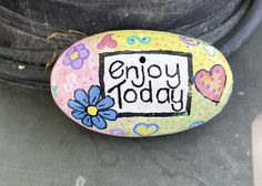 a painted rock that says enjoy today with flowers and hearts on it next to a tire