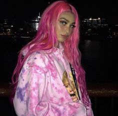 a woman with pink hair and blue eyes is standing by the water at night wearing a pink tie dye hoodie