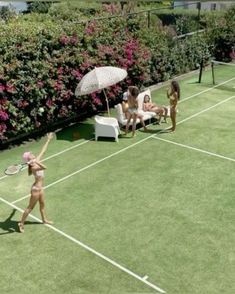 two women in bikinis playing tennis on a grass court with chairs and an umbrella