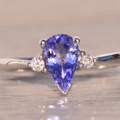 The 136th Avenue: Pear Shaped Tanzanite And Natural Diamond Ring In White Gold. The Ring Features A 1.00 Carat Pear Brilliant Cut Natural Tanzanite In The Center With A Medium Very Slightly Bluish-Purple Color. On Either Side Of The Center Is A Round Brilliant Cut Natural Diamond. A Tapered Shank Completes The Look. The Ring Is Crafted In 14 Karat White Gold And Is Currently A Finger Size 6 Yet Can Be Adjusted To Any Finger Size For An Additional Charge On Request. Purple Tanzanite Engagement Ring, Purple Diamond Ring, Tanzanite Rings, Crystal Healing Chart, Tanzanite Engagement Ring, Bluish Purple, Purple Diamond, Natural Diamond Ring, Tanzanite Ring