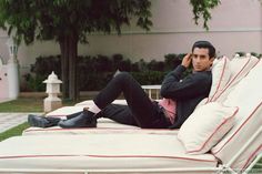 a man sitting on top of a white bed in front of a pink building with trees