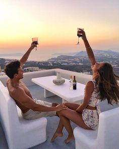 a man and woman sitting on top of a white couch holding wine glasses in the air