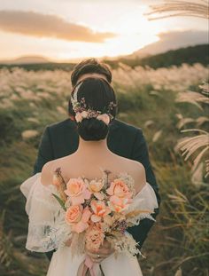 a bride and groom standing in tall grass with flowers on their head, looking into the distance