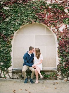 an engaged couple sitting on a bench in front of a wall covered with ivy