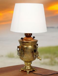 an antique brass urn lamp on a table in front of the ocean with a white shade