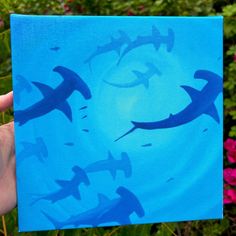 a hand holding up a blue painting with sharks in the water and pink flowers behind it