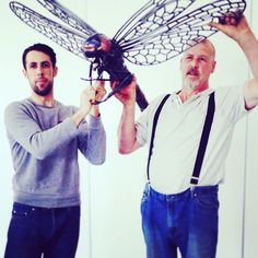 two men standing next to each other while holding up a model of a dragonfly