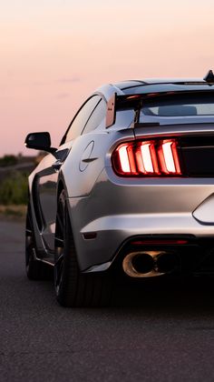 the rear end of a silver sports car driving down a road at dusk with its tail lights on