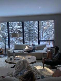 two people sitting on couches in front of large windows with snow covered trees outside