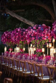 a long table covered in lots of purple flowers and lit candles next to a tree