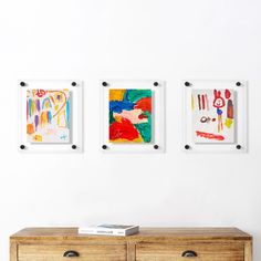 three framed art pieces hang on the wall above a wooden dresser in a white room