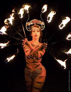 a woman with fire sticks in her hands and wearing a headdress, is surrounded by flames