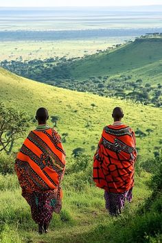 two people walking down a trail in the middle of an open field with green hills behind them