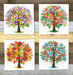 four cross stitch coasters with colorful tree designs on them, hanging on a wooden fence