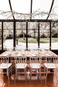 a dining room table set with white chairs in front of large glass windows and trees outside