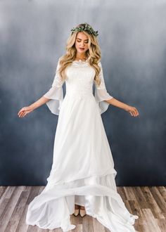 a woman wearing a white dress with sleeves and a flower crown on her head standing in front of a wall