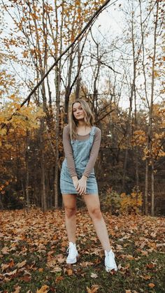 a woman standing in front of trees with leaves on the ground and looking at the camera