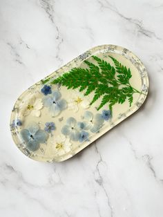 a tray with flowers and green leaves on it sitting on a marble counter top next to a plant