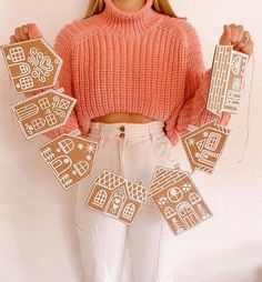 a woman wearing white pants and an orange sweater holds up some cut out gingerbreads