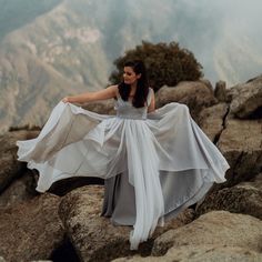 a woman in a white dress standing on top of a rocky hill with her arms spread out