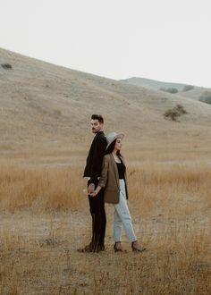 a man and woman standing in the middle of an open field with hills behind them