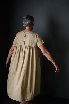 an older woman in a tan dress standing against a black wall with her hands behind her back to the camera