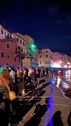 many people are standing on the sidewalk near the water and buildings at night with bright lights