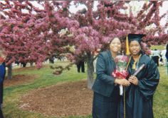 two women in graduation gowns pose for a photo under a tree with purple flowers