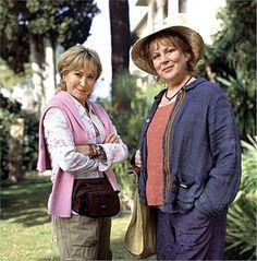 two women standing next to each other in front of some trees and bushes with their arms crossed