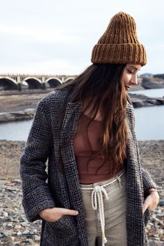 a woman standing on the beach wearing a jacket and hat with her hands in her pockets