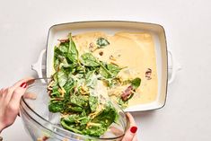 two hands holding a glass bowl filled with green salad and dressing on top of a white table