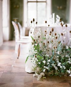 the table is set with white linens and flowers on it, along with clear chairs