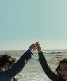 two people holding hands while standing in front of the ocean with their arms up to each other