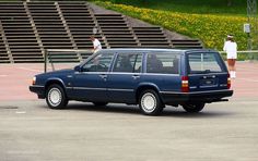 a blue station wagon is parked in front of some bleachers with people standing on the bleachers