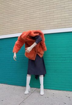 a mannequin dressed in an orange fur coat and white boots stands next to a blue brick wall