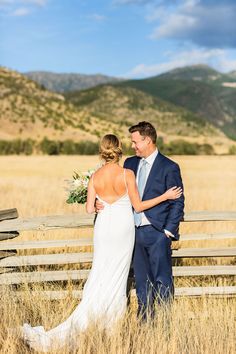 a bride and groom standing next to each other in front of a wooden fence with mountains in the background