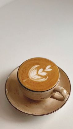 a cappuccino on a saucer sitting on a white counter top with a leaf drawn in the foam