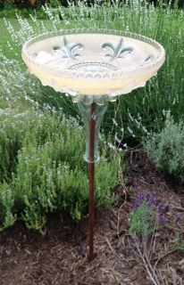 a bird bath sitting on top of a metal pole in front of some bushes and flowers